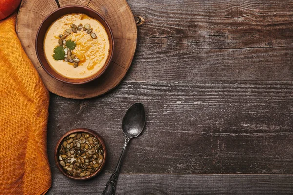 Vista dall'alto di ciotola con gustosa zuppa di zucca, cucchiaio e semi di zucca su tavolo di legno — Foto stock
