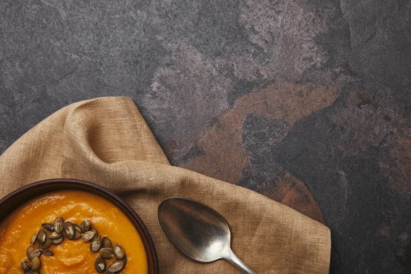 Верхний вид вкусного тыквенного супа с семенами и ложкой на мешковину на темной поверхности — стоковое фото