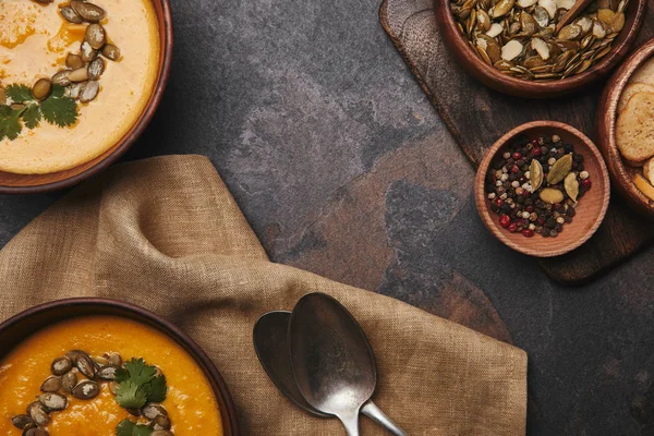 Vista dall'alto di deliziosa zuppa di zucca in ciotole, cucchiai su sacco e spezie con semi di zucca e fette biscottate sulla superficie scura — Foto stock