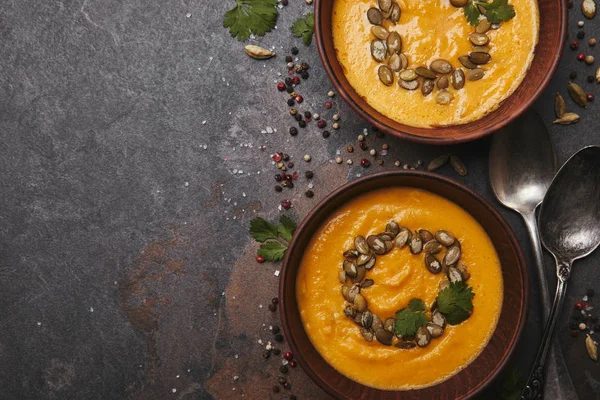 Vista dall'alto di gustosa zuppa di zucca sana con semi in ciotole e cucchiai su superficie scura — Foto stock