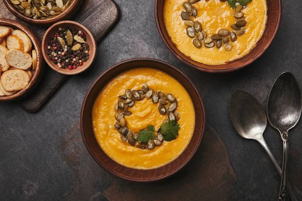 Vista dall'alto di ciotole con gustosa zuppa di zucca, cucchiai, spezie e fette biscottate su superficie scura — Foto stock