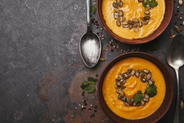 Vista dall'alto di zuppa di zucca sana con semi in ciotole e cucchiai su superficie scura — Foto stock