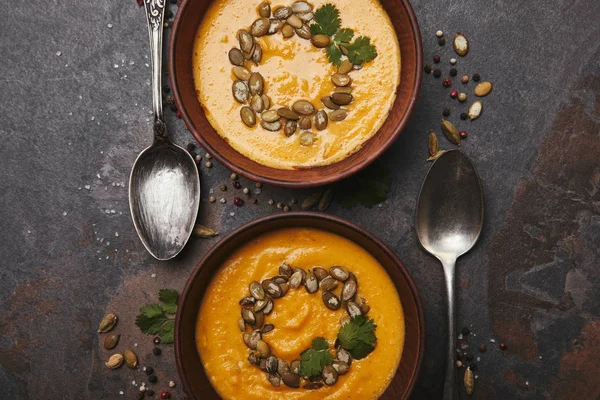 Vista dall'alto di ciotole con gustosa zuppa di zucca, cucchiai e spezie sulla superficie scura — Foto stock