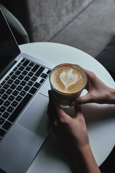 Partial view of person holding cup with cappuccino and using laptop — Stock Photo