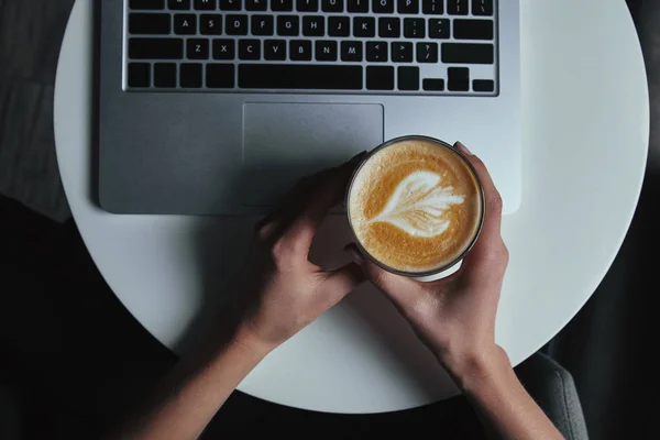 Tiro cortado de pessoa segurando copo com cappuccino e usando laptop — Fotografia de Stock