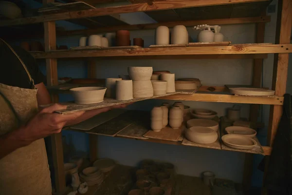 Immagine ritagliata di vasaio in grembiule mettendo ciotole di ceramica e piatti sugli scaffali in officina — Foto stock