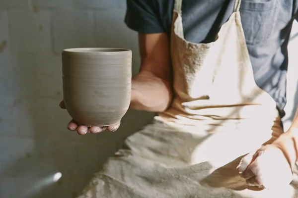 Imagen recortada de alfarero profesional en delantal sosteniendo olla de arcilla en el taller — Stock Photo