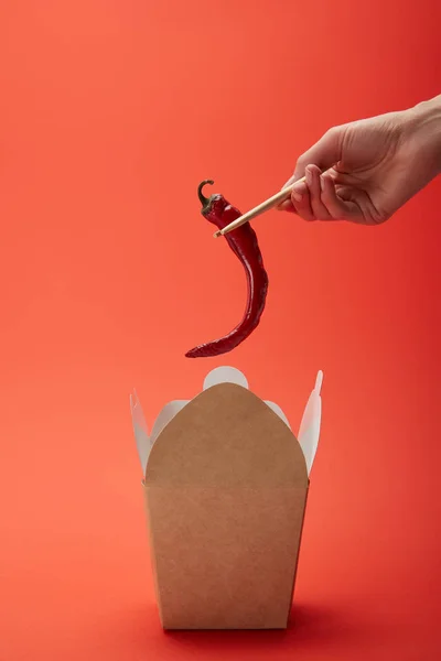 Cropped image of woman holding chili pepper in chopsticks above noodle box on red — Stock Photo