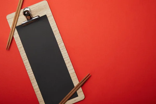 Blick von oben auf leeres schwarzes Menü und Essstäbchen auf roter Oberfläche, minimalistisches Konzept — Stockfoto