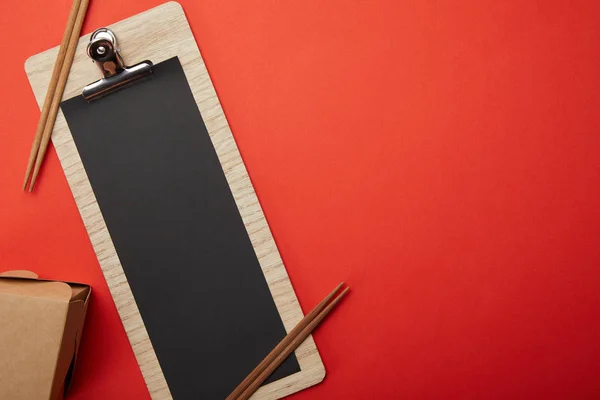 Top view of noodle box, chopsticks and blank black menu on red surface — Stock Photo