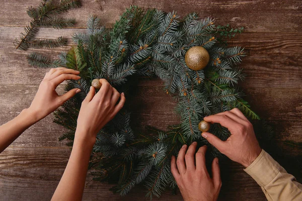 Imagen recortada de novia y novio decorando la corona de abeto de Navidad juntos en la mesa de madera - foto de stock