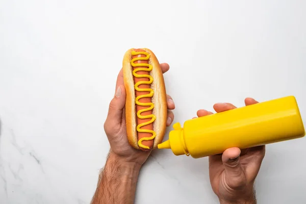 Colpo ritagliato di uomo versando senape su hot dog sulla superficie di marmo bianco — Foto stock