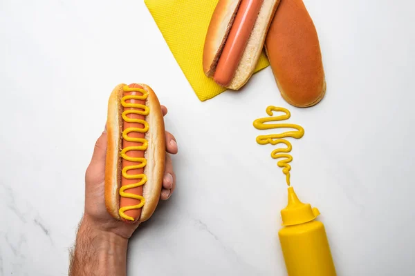 Cropped shot of man holding hot dog with mustard on white marble surface — Stock Photo