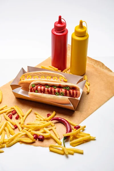 Close-up de cachorros-quentes picantes com batatas fritas, mostarda e ketchup em papel na superfície branca — Fotografia de Stock