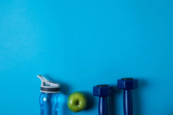 Elevated view of arranged sport bottle, green apple and dumbells isolated on blue, minimalistic concept — Stock Photo