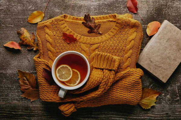 Acostado plano con hojas caídas, taza de té con trozos de limón, libro y suéter naranja en la mesa de madera - foto de stock