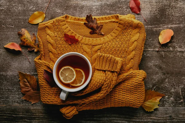 Acostado plano con hojas caídas, taza de té con trozos de limón y suéter naranja en la mesa de madera - foto de stock