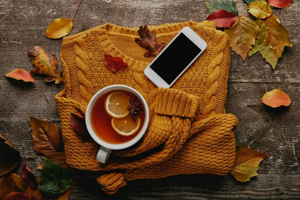Plat avec tasse de thé, pull ornement, smartphone avec écran blanc et feuilles tombées sur la surface en bois — Photo de stock