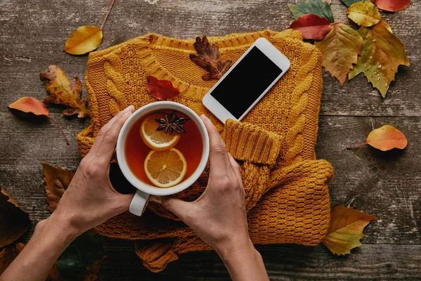 Vista parcial de la mujer sosteniendo la taza de té en la mesa de madera con suéter de punto, teléfono inteligente y hojas caídas - foto de stock