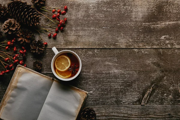 Flache Liege mit roten Stechpalmen, blankem Notizbuch, Tasse Tee und Tannenzapfen auf Holztischplatte — Stockfoto