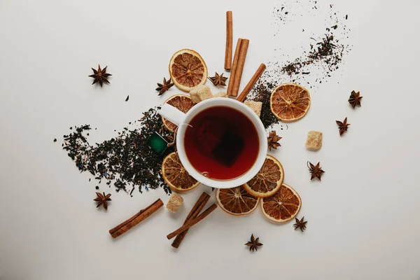 Tendido plano con palos de canela arreglados, estrellas de anís, piezas de naranja secas y taza de té caliente sobre fondo blanco - foto de stock