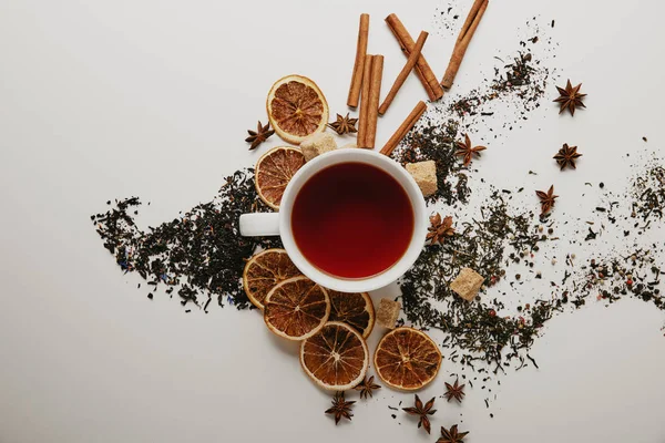 Plat étendu avec des bâtons de cannelle disposés, étoiles d'anis, sucre brun, morceaux d'orange séchés et tasse de thé chaud sur fond blanc — Photo de stock