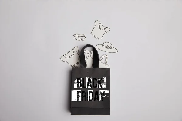 Top view of black shopping bag with black friday inscription and paper clothes on grey background — Stock Photo