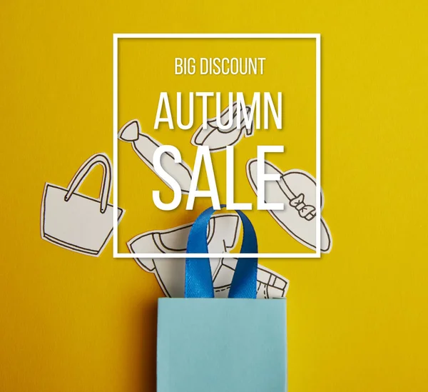 Vue du dessus du sac à provisions bleu avec des vêtements en papier sur fond jaune, inscription vente automne — Photo de stock