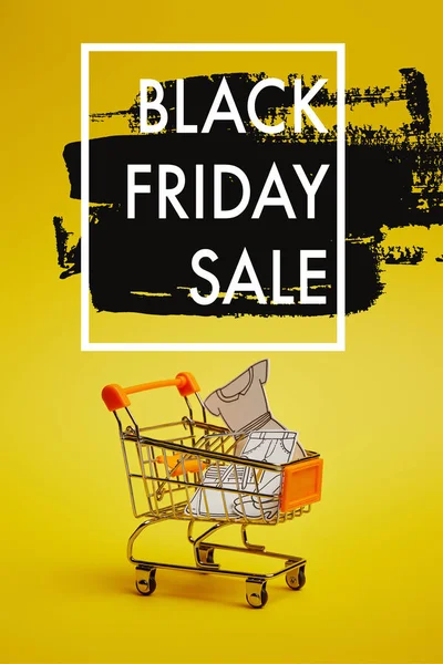 Крупним планом маленький торговий візок з паперовим одягом на жовтому тлі, чорна п'ятниця продаж напис — стокове фото