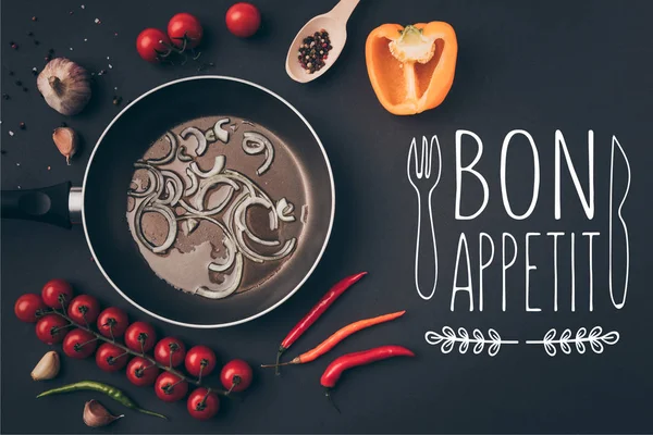 Top view of frying pan with onion and oil among vegetables on gray table, bon appetit lettering — Stock Photo