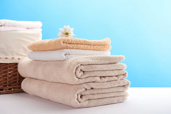 Pilha de toalhas macias limpas, flor de camomila e cesta de lavanderia em azul — Fotografia de Stock