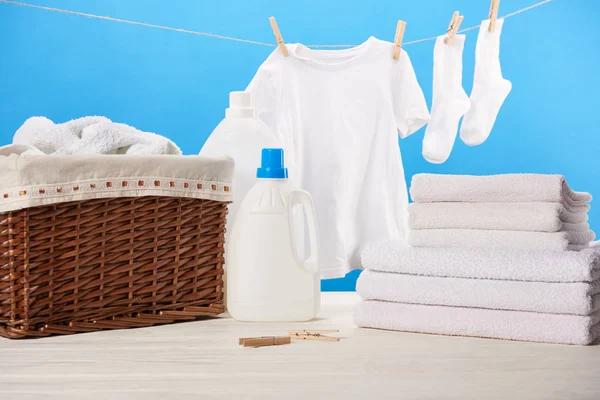 Récipients en plastique avec des liquides de lessive, panier à linge, pile de serviettes et des vêtements blancs propres sur bleu — Photo de stock