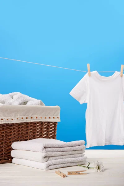 Panier à linge, pile de serviettes propres, pinces à linge, fleur de camomille et t-shirt blanc accroché sur corde à linge sur bleu — Photo de stock