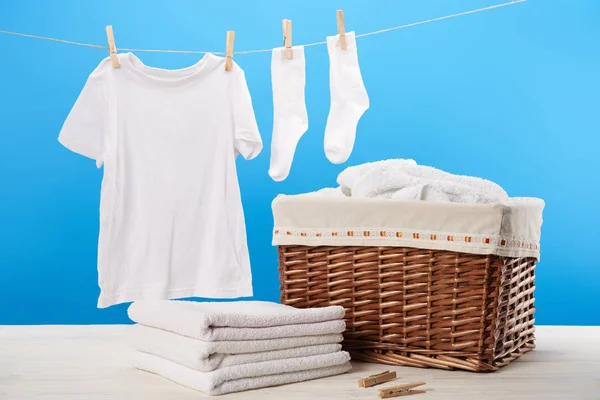 Panier à linge, pile de serviettes propres et des vêtements blancs suspendus sur corde à linge sur bleu — Photo de stock