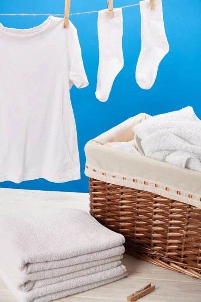 Vista de cerca de la cesta de la ropa, pila de toallas suaves limpias y ropa blanca colgando en el tendedero en azul - foto de stock