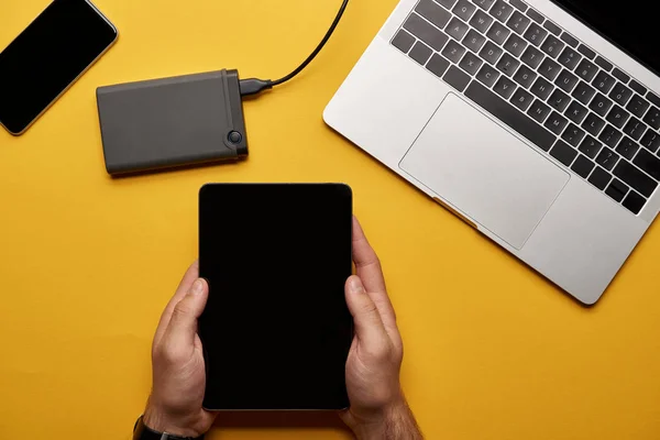 Обрезанный снимок человека с помощью планшета на желтой поверхности с ноутбуком и портативным hdd — стоковое фото