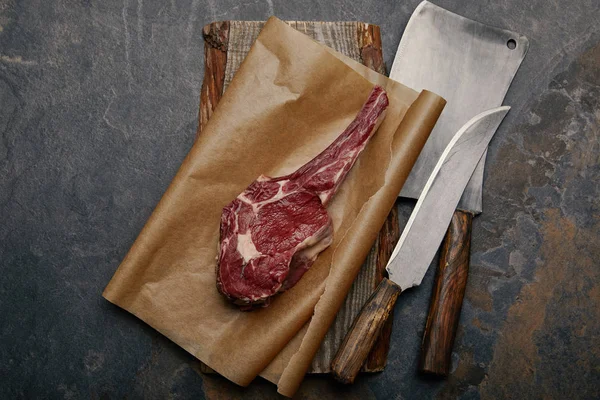 Vista dall'alto di costata cruda bistecca su carta da forno con coltelli da cucina su sfondo grigio — Foto stock