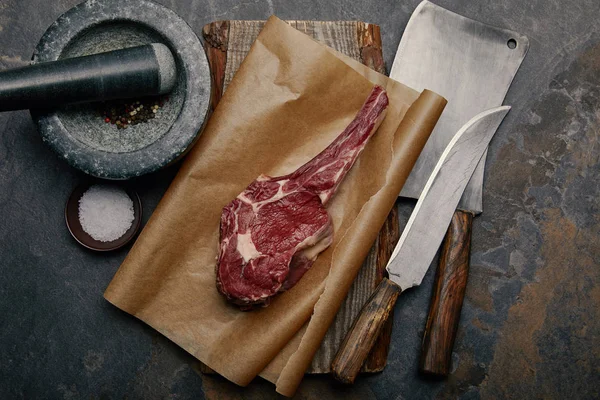 Top view of raw rib eye steak on baking paper with kitchen knives and marble pounder on grey background — Stock Photo