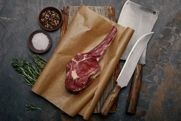 Vue du dessus du steak cru aux côtes levées sur papier cuisson avec couteaux de cuisine, épices, herbes et sel sur fond gris — Photo de stock