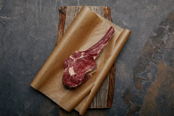 Top view of raw rib eye steak on baking paper and wooden board on grey background — Stock Photo