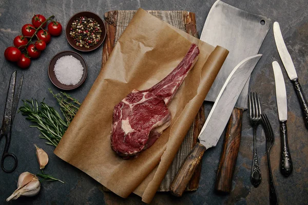 Ansicht von Roh-Rib-Eye-Steak auf Backpapier mit verschiedenen Küchenmessern, Gewürzen, Kräutern und Tomaten auf grauem Hintergrund — Stockfoto