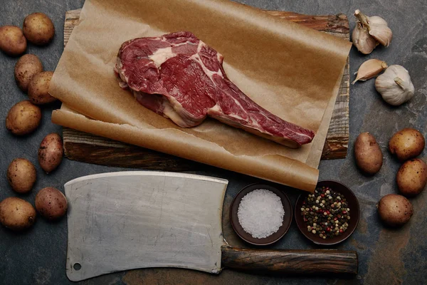 Flache Lage mit rohem Rippenaugensteak auf Backpapier mit Fleischermesser, Gewürzen, Kartoffeln und Knoblauch — Stockfoto