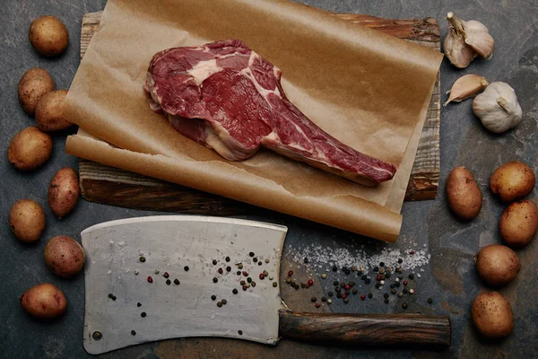 Tendido plano con filete de costilla cruda sobre papel de hornear con cuchillo de carnicero, especias, patatas y ajo - foto de stock