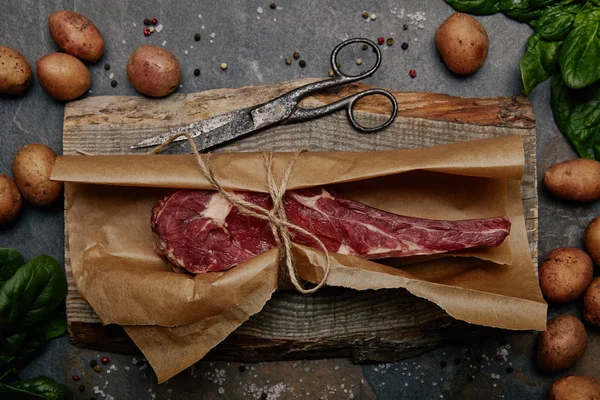 Top view of raw rib eye steak wrapped in baking paper on wooden board with spices and potatoes — Stock Photo