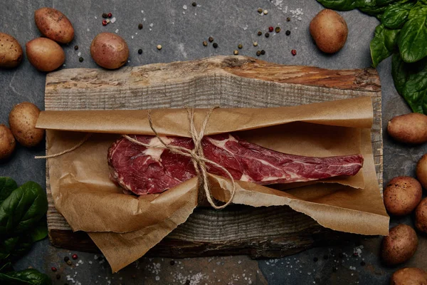 Bife de costela cru envolto em papel manteiga em tábua de madeira com especiarias e batatas — Fotografia de Stock