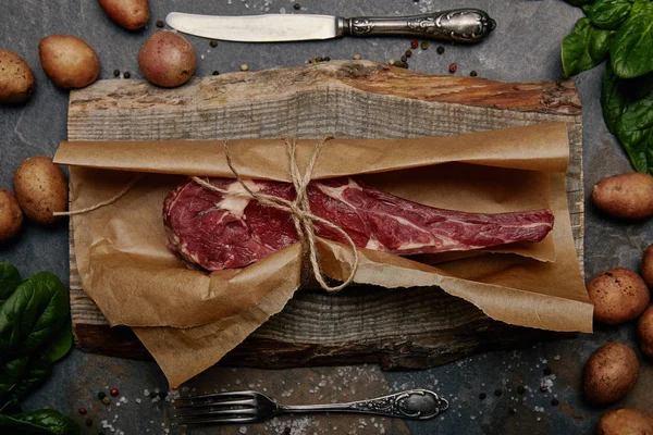 Bifteck aux côtes crues enveloppé dans du papier cuisson sur du carton en bois avec des épices, des pommes de terre et des couverts de cuisine — Photo de stock