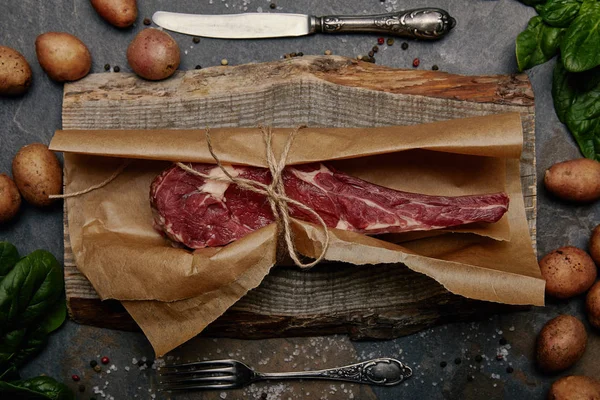 Bife de costela cru envolto em papel manteiga em tábua de madeira com especiarias, batatas e talheres de cozinha — Fotografia de Stock