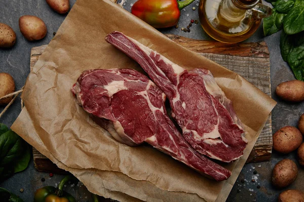 Raw rib eye steaks on baking paper on wooden board with potatoes and olive oil — Stock Photo