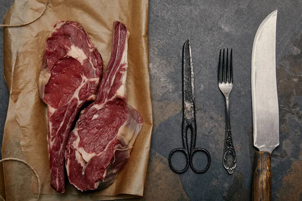 Vue du dessus des biftecks de côtes crues sur papier cuisson avec fourchette, couteau et ciseaux sur fond gris — Photo de stock