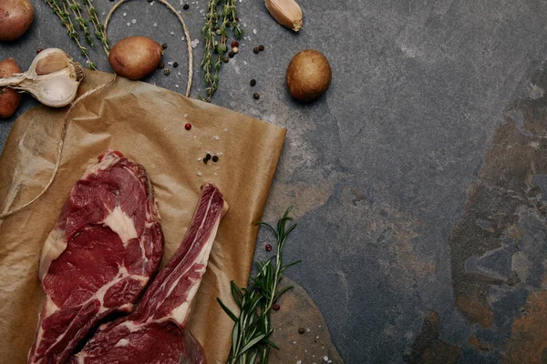 Tendido plano con filetes de costilla cruda sobre papel de hornear con especias, hierbas y patatas sobre fondo gris - foto de stock